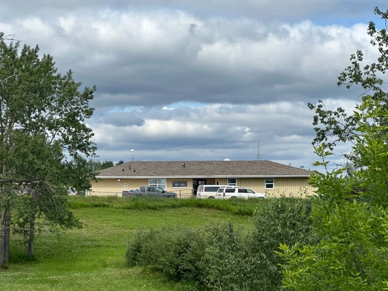 A pale yellow building is shown in the distance, past some trees. Several trucks and SUVs can be seen in front of the building.