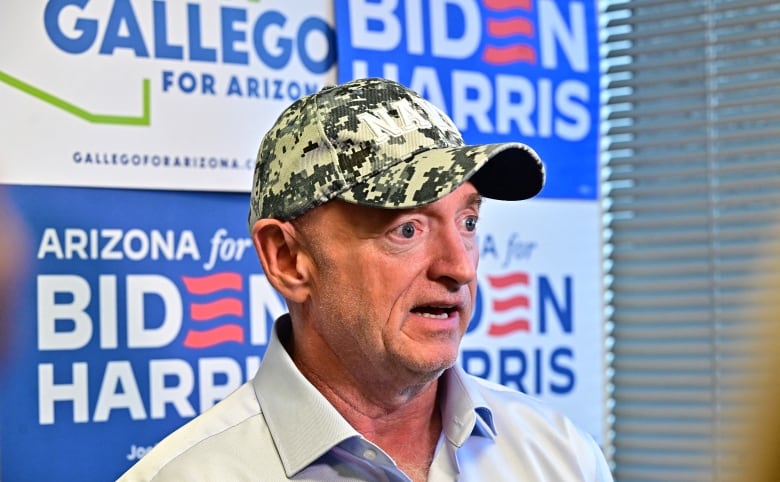 A man with a camouflage baseball cap is shown speaking in closeup in front of a wall that has political campaign signs.