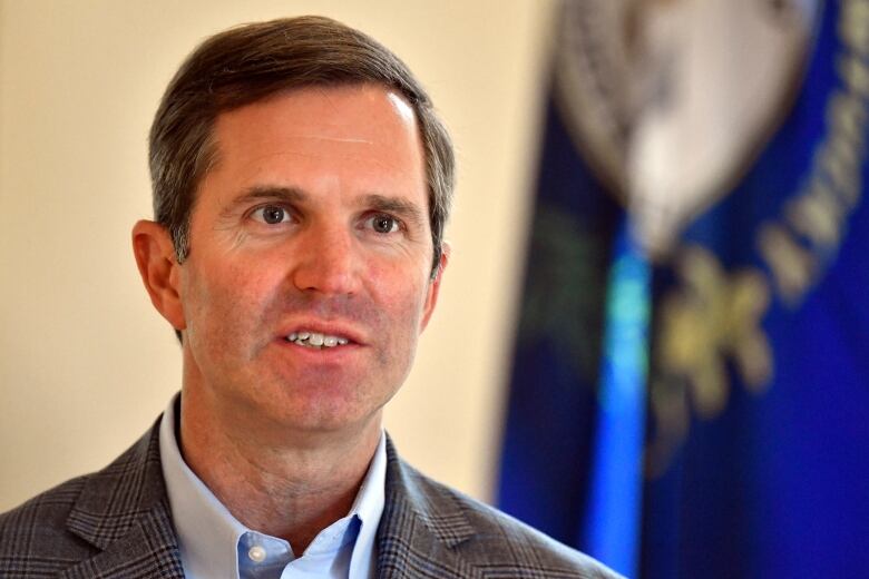 A cleanshaven man in a suit jacket and collared shirt is shown speaking in a closeup photo.