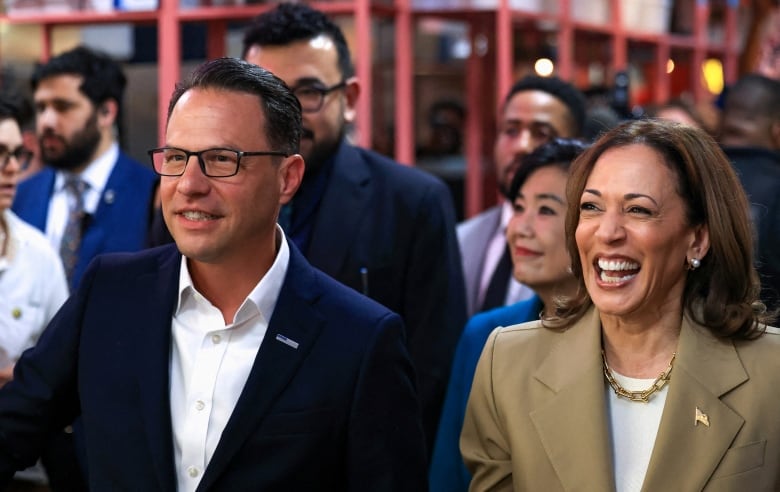 A bespectacled clean shaven man and a woman, both wearing blazers, smile during an indoor event.