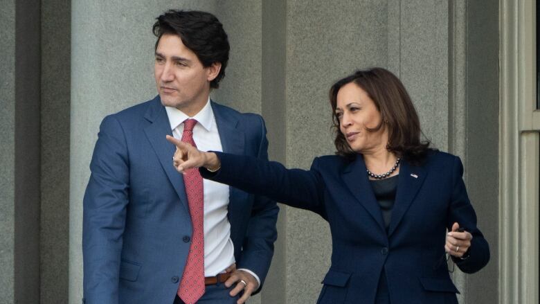 Justin Trudeau shown wearing a blue suit with a red tie standing on the White House balcony as Kamala Harris, who is wearing a dark blue suit and a black top, points into the distance. 