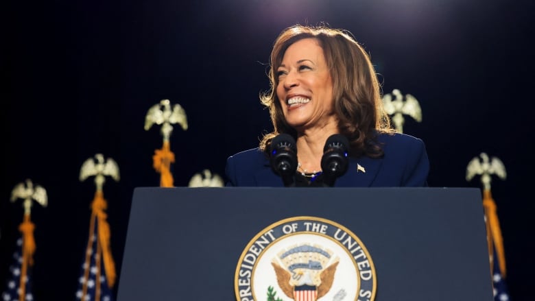 A woman in a blue suit stands at a podium with the vice presidential seal. American flags are behind her.