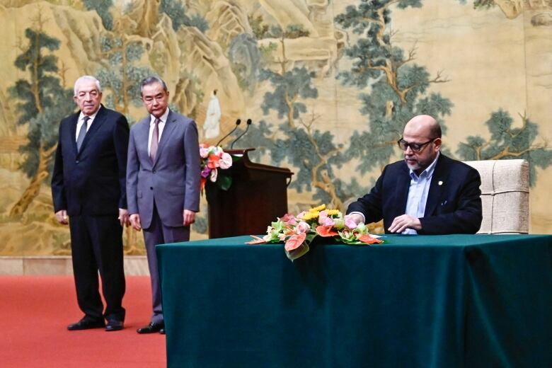 A man sits at a table to sign papers while two men watch him from a distance