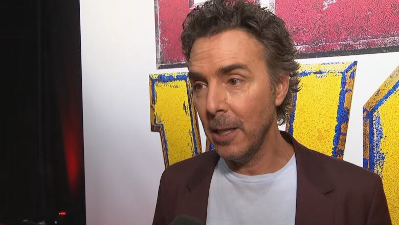 Shawn Levy wears a burgandy jacket and white shirt speaking to reporters on a red carpet