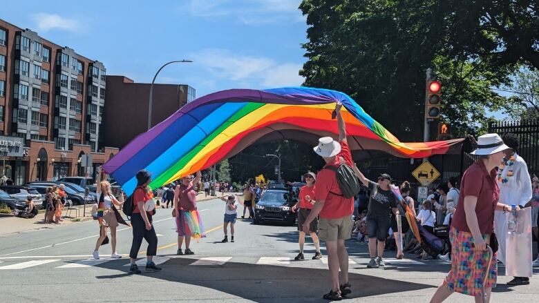 People at an intersection walking in the 2024 Halifax Pride parade hold a large rainbow flag. The flag was lifted and is flowing in the air.