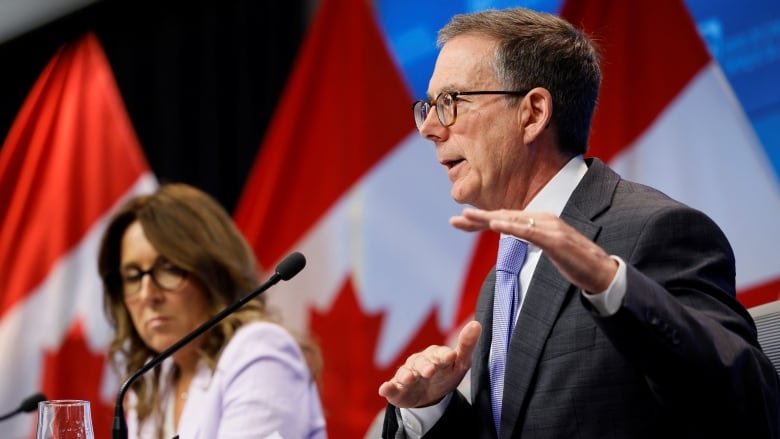 A man gestures while speaking during a news conference and a woman is shown seated to his right.