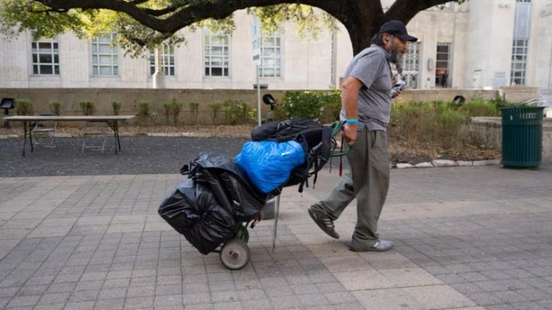 A man in a grey shirt pulls a trolley with some bags of his stuff.