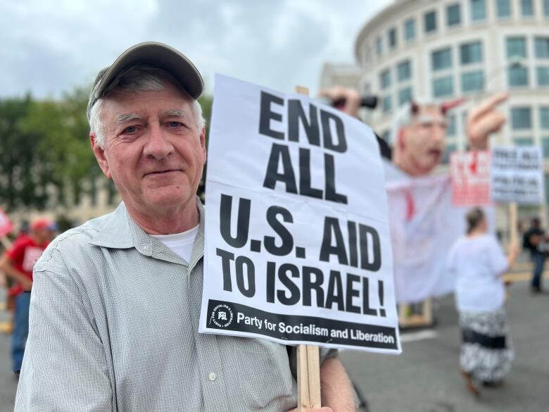 An older man in a button-down T-shirt and ball cap holds a sign that says 