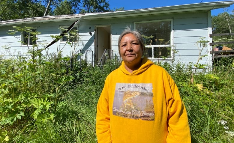 A woman stands outside a house where the weeds are high. The house looks deserted.