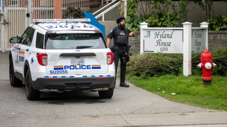 A Sikh police officer stands next to a police cruiser, outside a building marked 'Hyland House'.