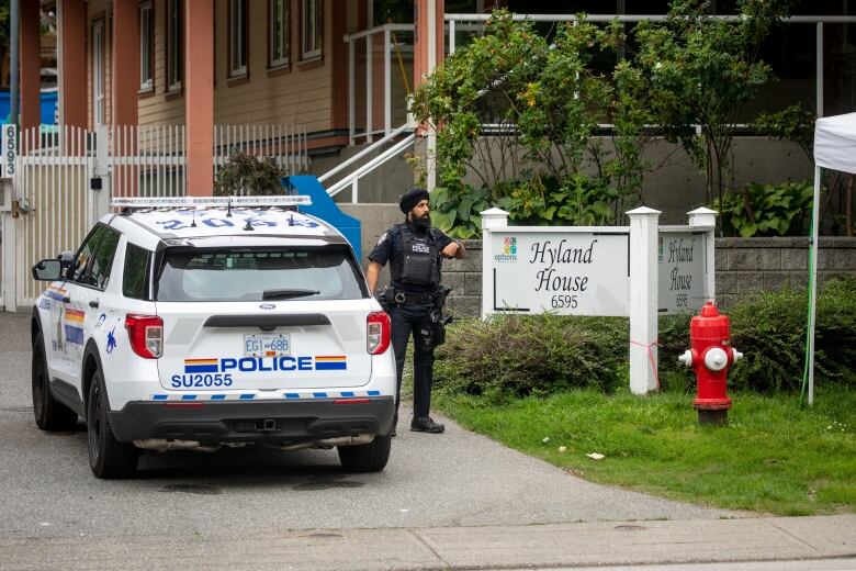 A Sikh police officer stands next to a police cruiser, outside a building marked 'Hyland House'.