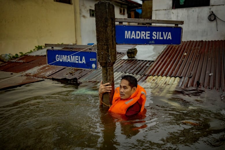 A man holds a street sign, wearing a life jacket, as water is up to this chest level.