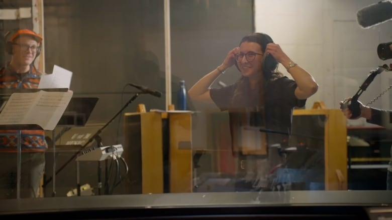 A woman wearing glasses and headphones smiles inside a recording booth. Another musician wearing an orange hat and glasses stands nearby.