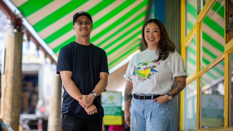 A man and a woman stand smiling outside in front of a market. 