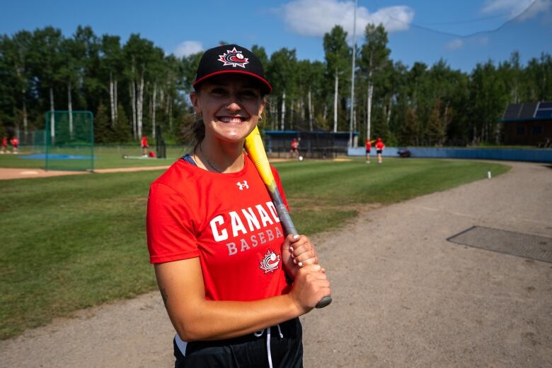 A woman in a red shirt holds a baseball bat and smiles for a photo.