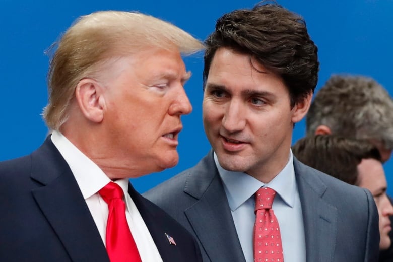 A man with gold hair wearing a navy suit and a red tie speaks while a younger man in a grey suit and red tie looks at him quizzically.