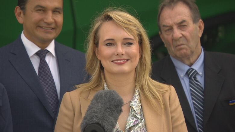 Two men in suits and a woman in a blazer stand behind a podium with a microphone.