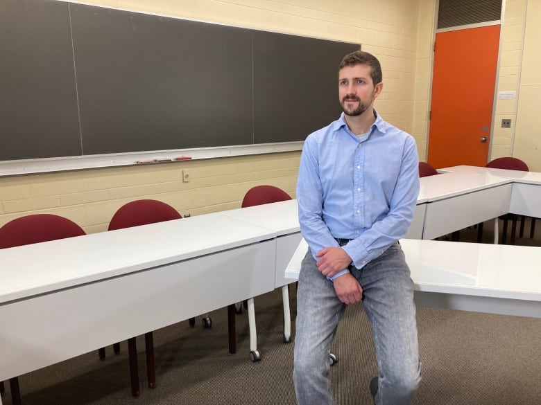 A white man in a blue shirt sitting on a desk in a classroom. 