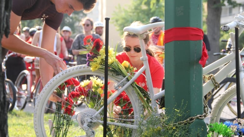 Two people place flowers around the spokes of a bicycle completely painted in white, which was chained to a green pole.