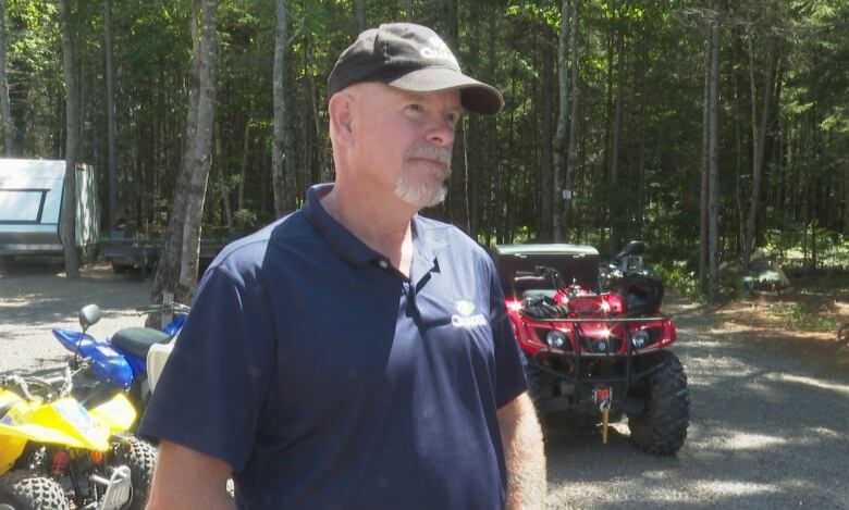 A man in a baseball cap and polo shirt, standing in front of a wooded area with several off-road vehicles in the background. 