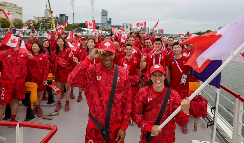 Two athletes, a man and a woman, are seen posing with the Canadian flag on a boat, in front of plenty of fellow athletes.