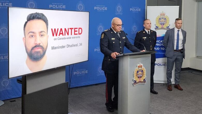 A TV screen displays the face of a man. A bald man in Edmonton police uniform stands before a podium. Two other men stand to his side, one in an RCMP uniform and one in a grey suit. Edmonton Police Service emblems can be seen on a backdrop.