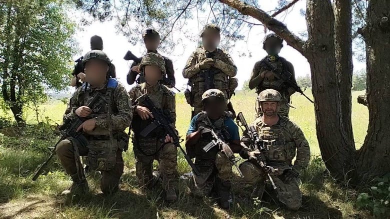 Eight soldiers pose for a photo in war gear. Seven of the faces are blurred.