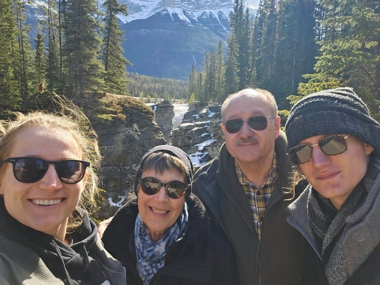 A family of four with a mother, father, daughter, and son stand in front of a frozen waterfall. There are evergreen trees on either side of the waterfall in the background, and the base of a mountain in the distant background.