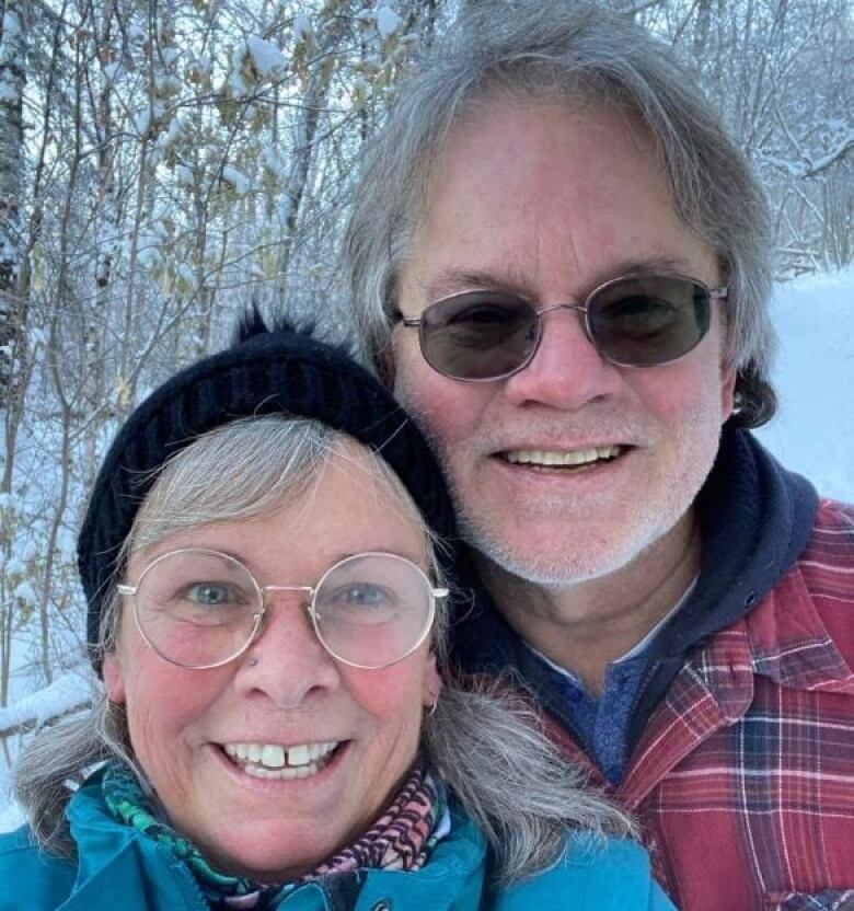 A woman in a blue coat with round glasses and a toque stands next to a man in a red flannel coat with sunglasses. In the background are snow covered trees.