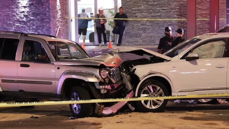 Two cars are pictured crashed into each other behind crime tape, while people look on.