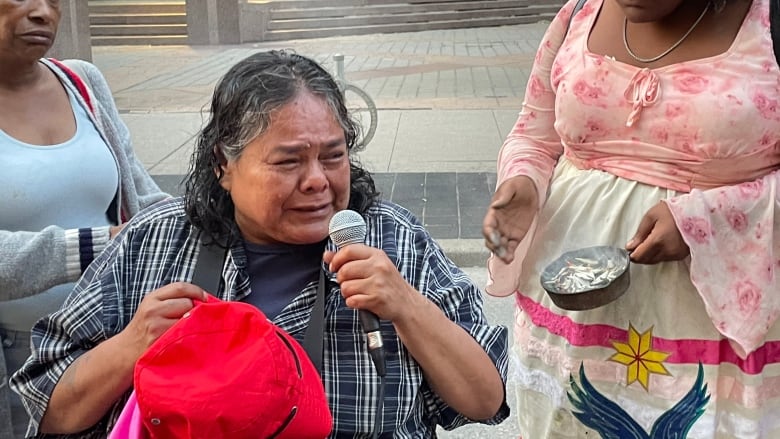 An elderly woman holds a microphone on a city street. She is in a wheelchair, crying. Two woman console her on either side of her. It is day time. 