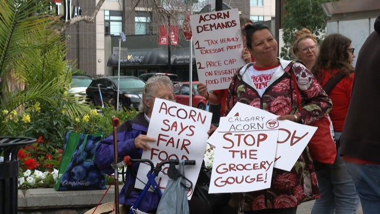 A group of people hold signs on 17 Avenue southwest. One person with grey hair wearing a blue jacket, whose face is obscured, holds a sign that reads 