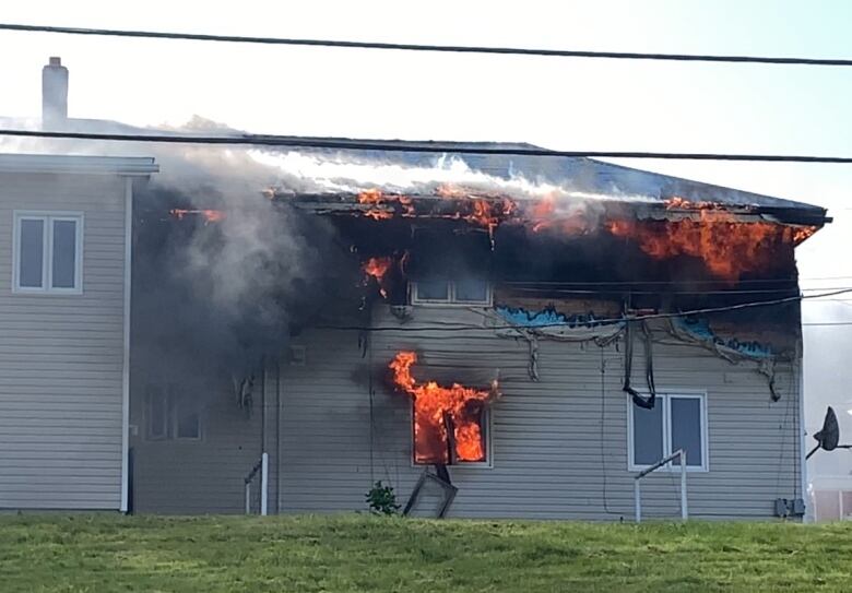 A housing complex is covered in flames. Vinyl siding is melting while flames flow underneath a roof. 