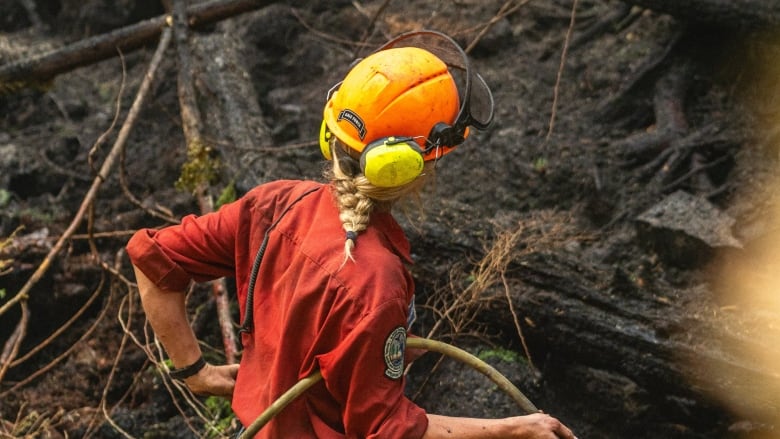 A woman in an orange shirt with a blond braid under a yellow hard hat holds a hose in a charred forest.