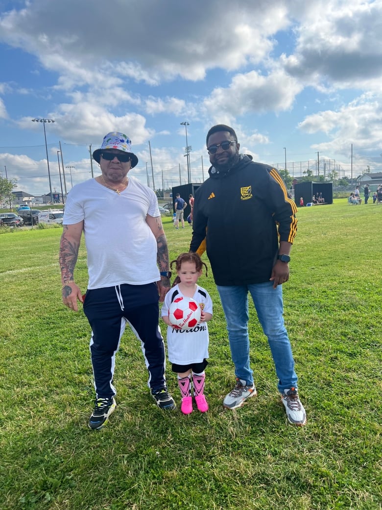 On a soccer field, the man on left wears white t-shirt, black pants, bucket hat and sunglasses. Man on left wears black Adidas jacket with yellow stripes, jeans and sunglasses. Standing in front of them is a little girl with pig-tails holding a soccer ball in pink cleats and shin pads and white t-shirt. 