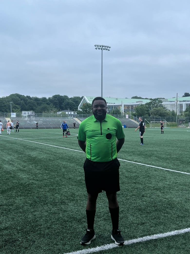 Man stands on grassy soccer field wearing black shorts, socks and sneakers and neon green referee jersey.