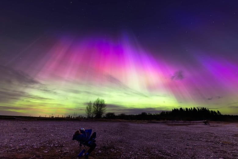 A person lies in a chair in the foreground while the sky glows a spectacular purple, pink and green.