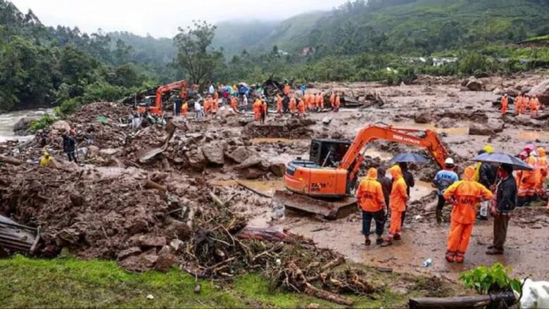 Dozens of people in bright reflective rain gear are shown from a distance on muddy ground, with some utilizing heavy machinery.