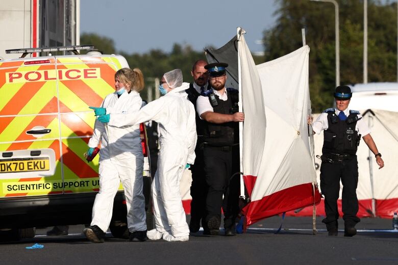 Two people in white suits and latex gloves stand near a police van with the words 