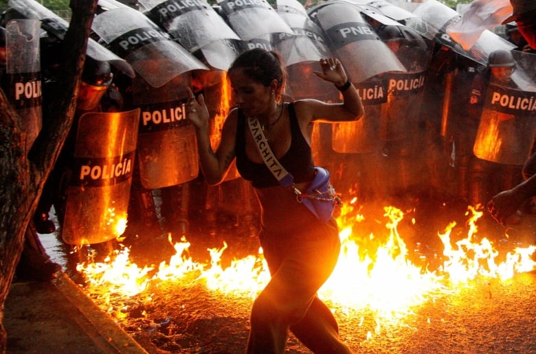 A woman runs with her hands up, fire on the ground behind her and a phalanx of police holding riot shields behind that.