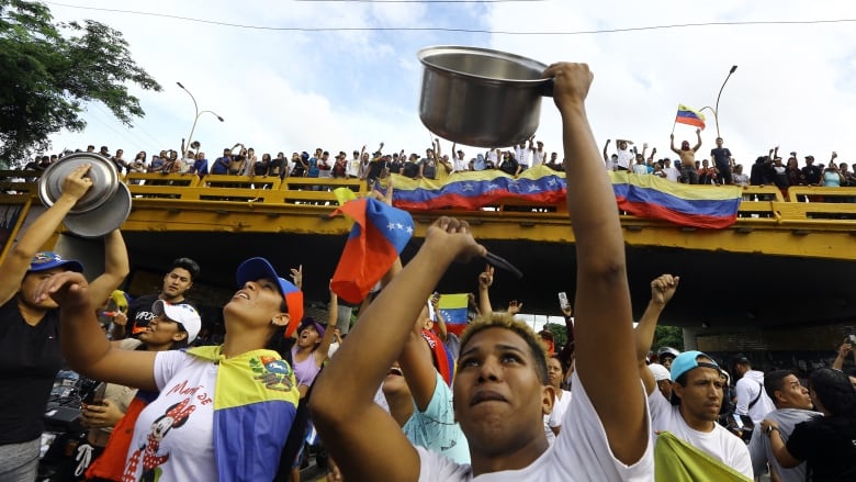 A crowd with many Venezuelan flags stands over and under an overpass. Two people clang pots.