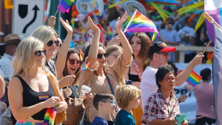 People cheering at a parade. They are wearing rainbow items of clothing and holding rainbow flags.