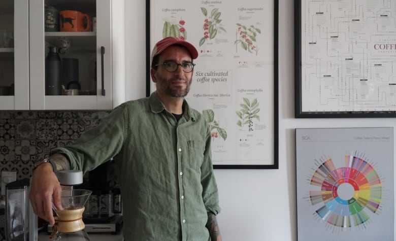 A man in an olive-green button-up and red cap and glasses leans against a specialty coffee machine. Behind him are several framed coffee posters.