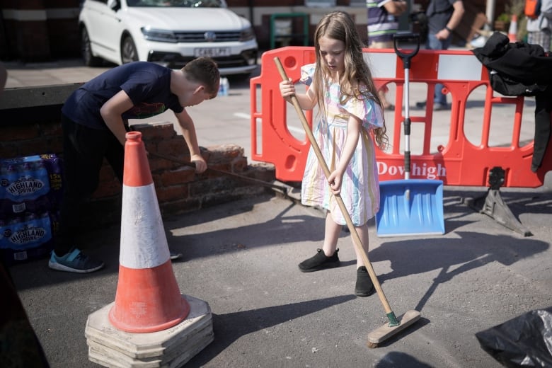 Two young children hold push brooms near an orange pylon.