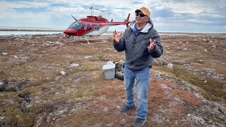 A man in a jacket, jeans, sunglasses and baseball cap on speaks in front of a helicopter.