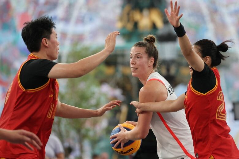 A female three by three basketball player controls the ball with both hands as two opponents defend with their arms in the air.