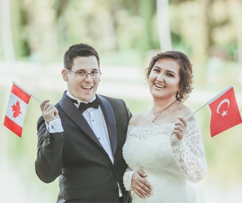 A woman in a wedding dress holds a Turkish flag. A man in a tuxedo hold a Canadian flag. 