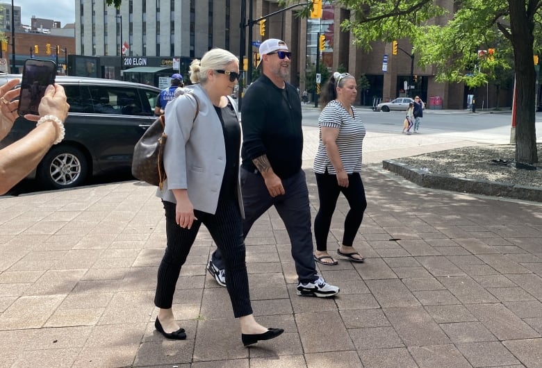 A man in a black sweater, white ballcap worn backwards, and sunglasses walks outside on a city street, surrounded by a few other people. 