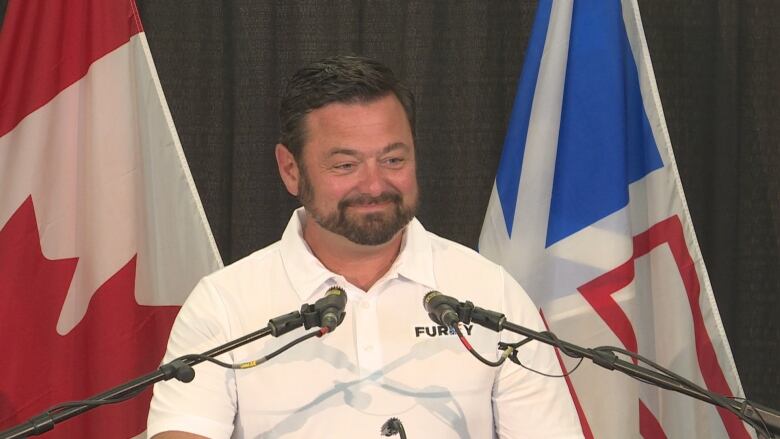 Man smiles with two mics in front of him and a Canadian and Newfoundland and Labrador flag behind him.