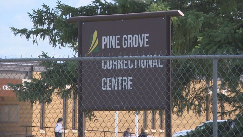 A chain-link fence is in the foreground with a Pine Grove Corrections Centre sign in the background behind the fence